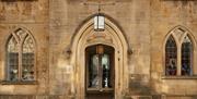 Arched entrance of an old stone building with gothic-style windows on either side and a hanging lantern above the doorway. Reflections and interior de