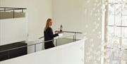 A woman in black clothing stands on an upper floor balcony, holding a tray with a wine bottle and glasses, looking back and smiling. Glass crystal dec