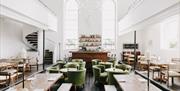Modern restaurant interior with high ceilings, large arched windows, green cushioned chairs, wooden tables, and a central bar; a spiral staircase is v