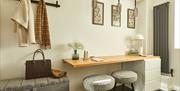 A wooden desk, two gray stools, wall-mounted hooks holding coats, a handbag on a bench, framed art on the wall, a white lamp, and a vase with flowers
