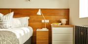 A white bed, gray and white pillows, a wooden headboard, white and wooden nightstands, a white lamp, a beige vase, yellow books, and sunlight streamin