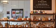 Shelves display a variety of fresh breads and pastries, each labeled with a small sign. Above, a chalkboard sign advertises soup of the day and chocol