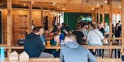 Families eating food while sat around wooden benches in a room with wooden beams and string lights
