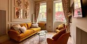 A warmly lit living room with large windows and floral full-length curtains, a wall-mounted TV, large yellow sofa and two armchairs.