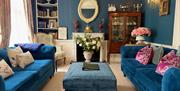 A living room with two blue sofas and an ottoman, a white fireplace and against a blue wall, and an ornate mirror.