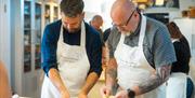 Two men in white aprons prepare food on a kitchen worktop.