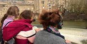 Three people wearing silent disco headphones face away from the camera towards Pulteney Weir in Bath.