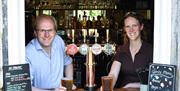 Two people holding pints over the bar at The Cross Guns pub in Avoncliff, near Bath.