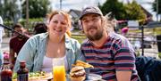 A man and woman smiling, sitting outside eating burgers and drinking orange juice