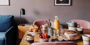 A dining table with a loaf of rustic bread, croissants, fruit, orange juice, a teapot and several mugs and glasses.