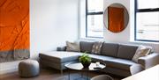 A living room with a large grey corner sofa, round coffee table and a red and grey textured art piece on the wall.