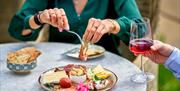 A person helping themselves from a cheeseboard while another person holds a glass of red wine at Iford Manor Kitchen.