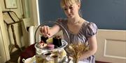 A woman in a purple regency-era dress reaches for a cake on an tiered afternoon tea platter.