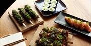 A wooden table with four plates of sushi and meat kebabs on square plates and wooden blocks.