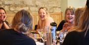 A group of women laughing around a dining table post-meal with wine glasses and a stone wall in the background.