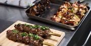 A metal cooking tray of kebabs on sticks next  to a serving wooden block with two of the kebabs on it.