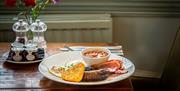 A plate containing a breakfast of fried egg, sausages, bacon, a hash brown, and baked beans in a ramekin.