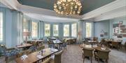 A horizontal view of the restaurant with soft chairs, wooden tables and a chandelier hanging above.