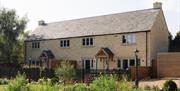 The outside of sandstone row of two cottages with a slate pitch roof and porched front doors with a brown picket fence outside and a garden full of wi