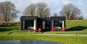 Modern-style black and white accommodation building with red chairs in front, overlooking a lake.