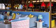 Blue, yellow and pink beach themed comfy sofas, chair and table in front of a bar that serves cocktails.