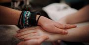 A pair of hands wearing a watch and several beaded bracelets massage a skincare product into another person's hand.
