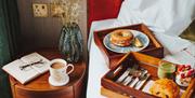 Two breakfast trays with a smoothie, pastry and a bagel on a bed edge, next to a bedside table with a coffee and open notebook.