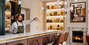 Bar tender pouring a cocktail on a white marble topped bar with a lit fireplace and bookshelf in the background.
