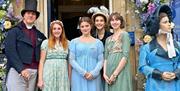 Five staff members in regency costumes pose in front of the entrance to the Jane Austen Centre, next to a statue of a regency-era woman.
