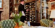 Waiter dressed in a black suit looking at wines in an ornate wall cellar/fridge in a restaurant.
