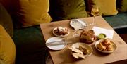 Several plates of food on a square table, next to a bench with green and yellow cushions