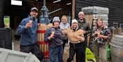 The team at Saddle Goose pose for the camera while smiling and holding wine glasses, surrounded by winery equipment at the warehouse.