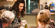 A lady with long dark hair and a decorative sleeved dress teaching a cookery class to three people.