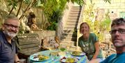 A group of three people sat around a sunny table in an attractive courtyard with white wine and plates of food.