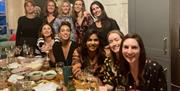 A group of 10 girls smiling and toasting along a long wooden table with lots of empty plates and wine glasses on.