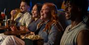 Four people sat in theatre chairs with drinks and popcorn, smiling and laughing at something out of shot.