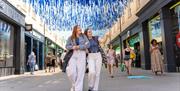 Two people stand in the middle of a high street with rows of shops either side. Lots of blue and white ribbons are hung overhead.