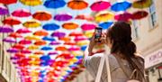 A woman holds a phone up to take a photo of rows of brightly coloured umbrellas hung above the high street