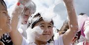 Children playing joyfully in a cloud of foam bubbles under a bright sky. A young girl in the center, smiling with arms raised, is covered in bubbles.