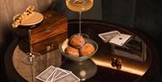 Two cocktails on a table top with a bowl of doughnuts and playing cards.