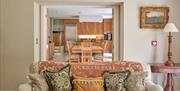 A grey sofa with ornate William-Morris style cushions on and an open plan kitchen diner with wooden table and chairs in the background through some do