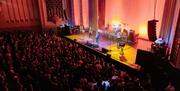 A busy crowd of combined seating and standing event attendees watch a band play live at The Forum, with red lighting and yellow spotlights.