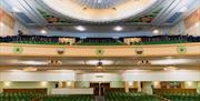 Theatre-style seating at The Forum, with many rows of green chairs. More rows of seating are visible on the balcony above. All seats are empty.