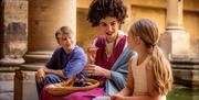 A woman dressed in costume as a Roman talking to two children next to the Great Bath at The Roman Baths.