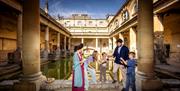 A woman dressed in costume as a Roman talking to a family of two adults and two children next to the Great Bath at The Roman Baths.
