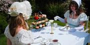 Two women dressed in Regency attire eating an alfresco afternoon tea in the garden of the Roseate Villa Bath
