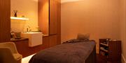 A spa treatment room with a bed with a grey towel and pillow on, a large mirror on the wall with white cuboid sink in front of it. Low level lighting.