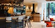 A smart indoor bar and seating area with white and black bar stools around a curved black panelled bar with white sofas to the right and a floor to ce