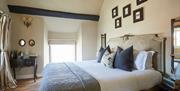 A bedrom with a a large bed with white sheets and various pillows. There is a small wooden desk in the corner and a window in the background