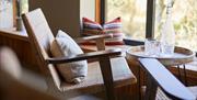 A chair with a pillow facing a table that holds a crystal glass and bottle in front of a window